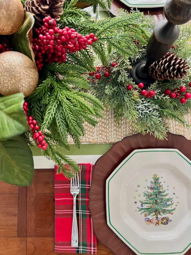 A festive table setting with a Christmas tree plate on a brown charger, a red plaid napkin with a fork, and a holiday centerpiece of evergreen branches, pinecones, gold ornaments, and red berries.
