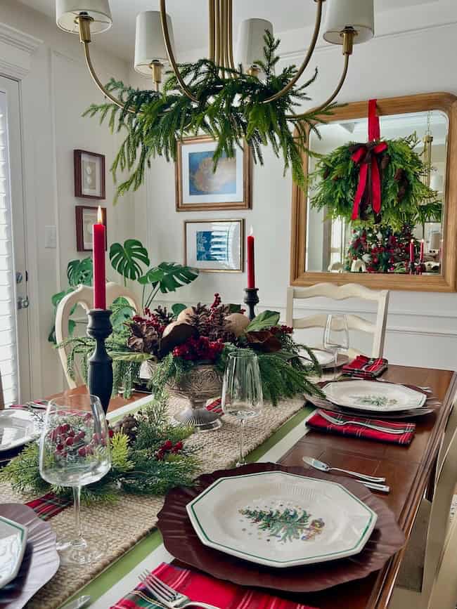 A festive dining table set with holiday plates, red candles, greenery, pinecones, and plaid napkins. A wreath with a red bow hangs on a mirror behind the table, and a chandelier is decorated with green branches.