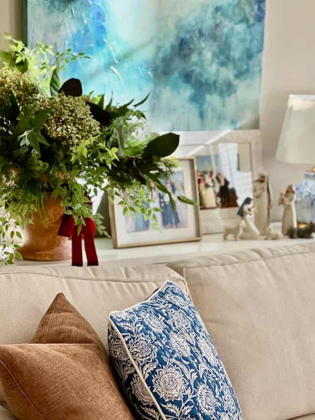 A beige sofa with brown and blue patterned pillows sits in front of a table holding framed photos, a lamp, porcelain figurines, and a large green floral arrangement. An abstract blue painting hangs on the wall.