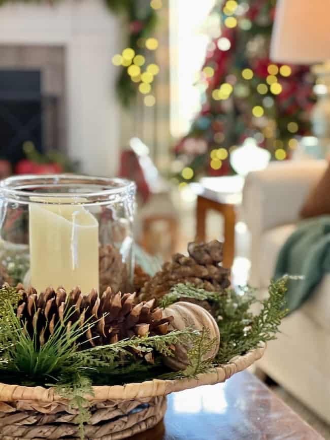 A woven tray holds a glass candle holder, a cream-colored candle, pinecones, greenery, and a wooden spool; a decorated Christmas tree and blurred lights are visible in the background.