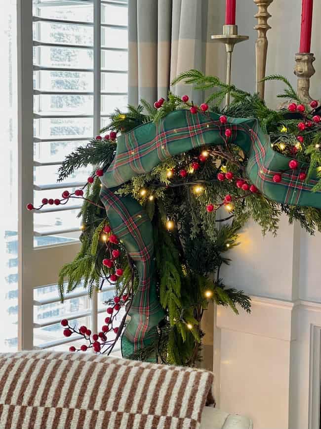 A festive Christmas garland with plaid ribbon, red berries, and twinkling lights decorates a mantel next to a window with white shutters. A striped blanket is draped over a nearby chair.