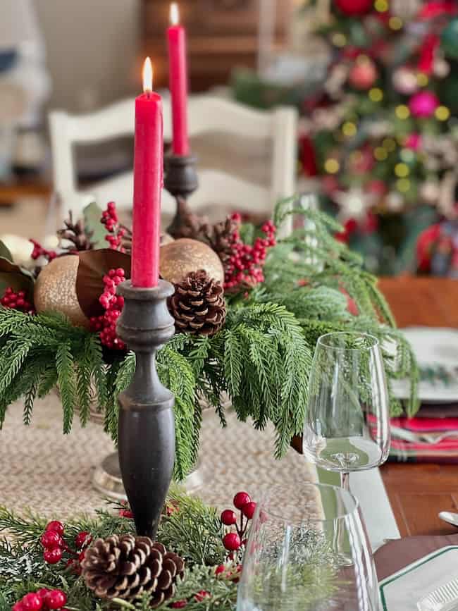 A festive table centerpiece with red candles, pinecones, red berries, and evergreen branches, with a blurred Christmas tree and dinnerware in the background.