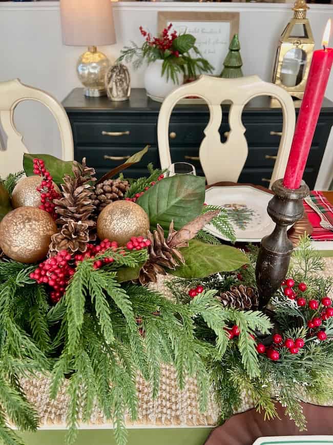 Festive table centerpiece with evergreen branches, red berries, pinecones, gold ornaments, and a red candle in a dark holder—perfect for a traditional plaid Christmas home tour. Cream chairs and holiday decor accent the background.