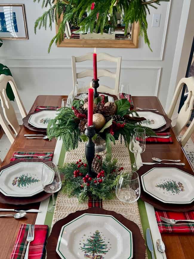 A dining table set for six with holiday-themed plates, red plaid napkins, wine glasses, and festive greenery centerpieces. A tall red candle sits in the middle, and a decorated wreath hangs above the table.