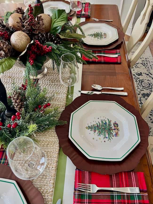 Festive holiday table setting with plates featuring Christmas trees, plaid napkins, wine glasses, and silverware. A centerpiece with pinecones, greenery, and red berries decorates the wooden table.
