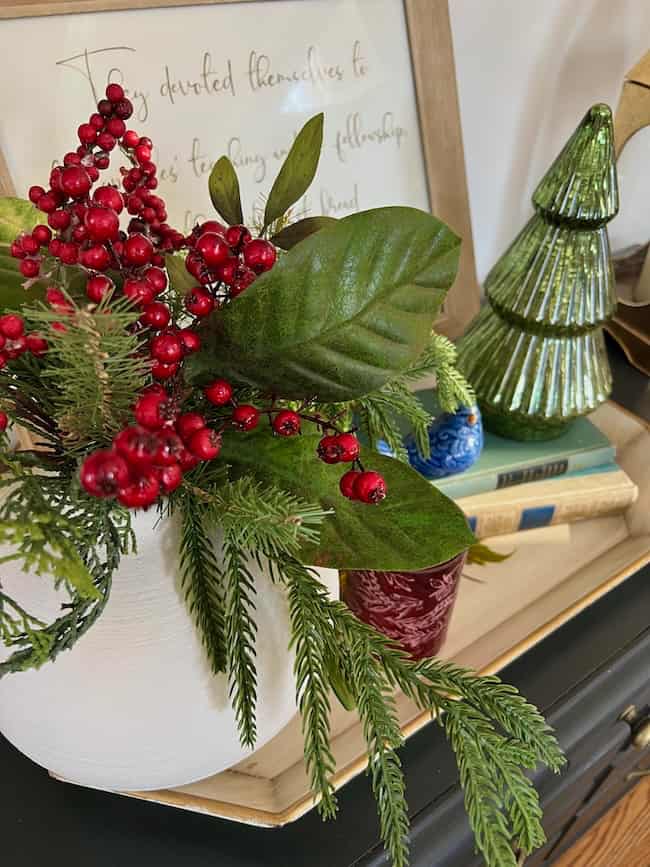 A white vase with green leaves, red berries, and pine branches sits on a tray near glass Christmas trees and stacked books, perfect for a traditional plaid Christmas home tour festive holiday display.