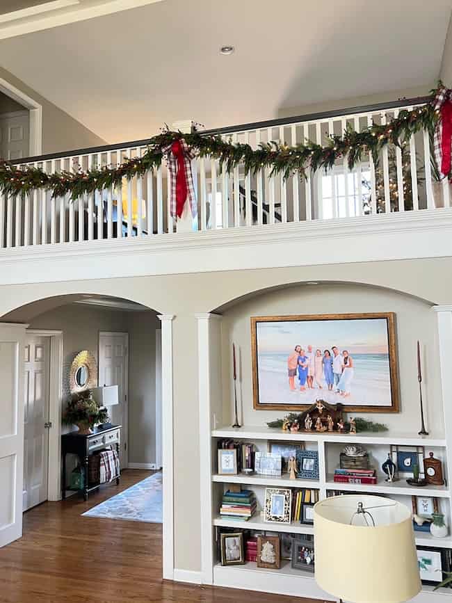A bright living room decorated for Christmas, featuring a garland with red ribbons on a balcony railing, built-in shelves with books and décor, a nativity scene, and a large family photo above the shelves.