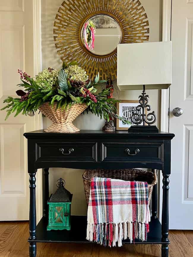 A black console table with drawers holds a basket of flowers, a decorative lamp, and framed art. Below, a woven basket with a plaid blanket and a green lantern sit on the lower shelf. A round gold mirror hangs above.