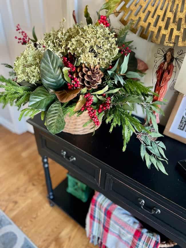 A decorative black table holds a festive arrangement of green leaves, red berries, dried hydrangeas, pinecones, and evergreens in a woven basket. A colorful throw blanket sits on the lower shelf.