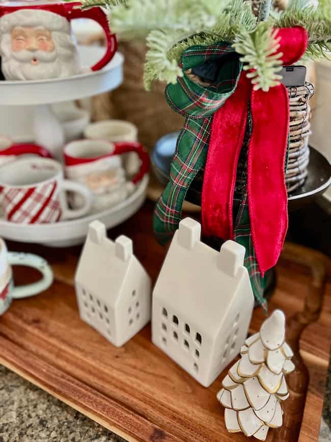Two white ceramic house-shaped decorations and a white Christmas tree ornament sit on a wooden tray beside a festive display of mugs, including a Santa mug, and a tiered tray with greenery and a red plaid ribbon.