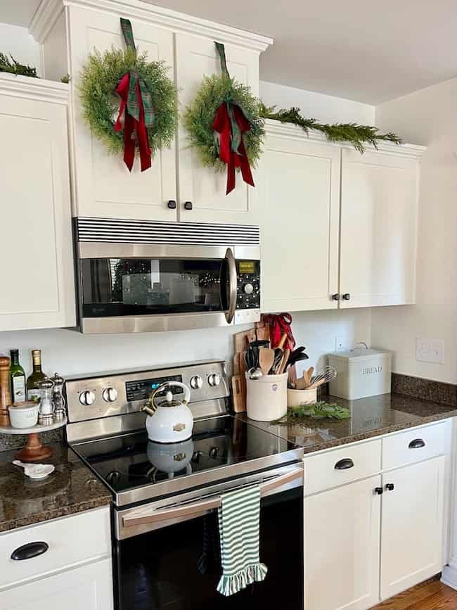 A modern kitchen with white cabinets decorated for the holidays; two green wreaths with red ribbons hang above a stainless steel stove and microwave, with greenery on top of the cabinets.