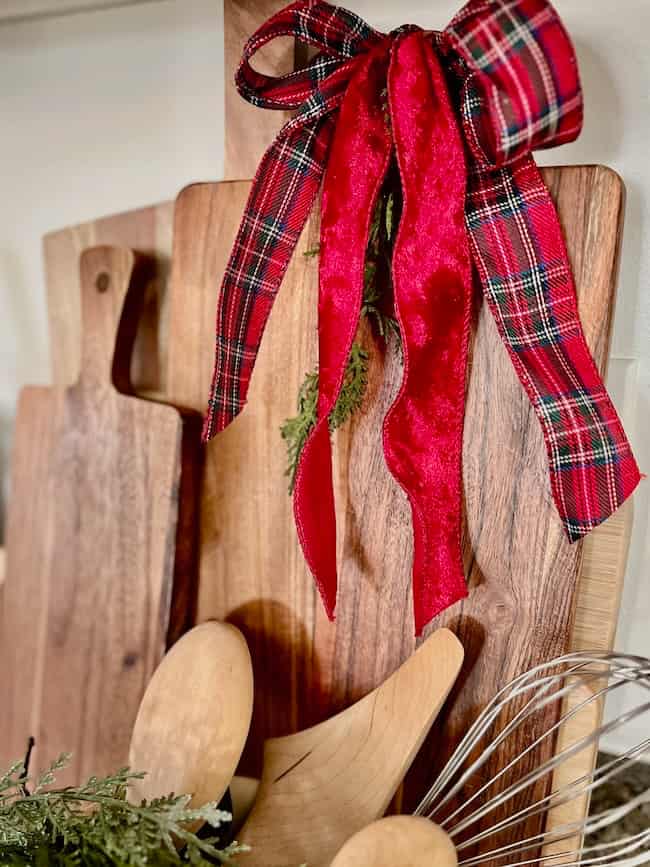 Several wooden cutting boards stand upright, with one decorated by a red plaid ribbon and some greenery. Wooden utensils and a metal whisk are placed in front of the boards.