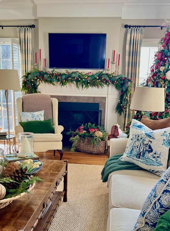 A cozy living room decorated for Christmas, featuring a fireplace with garland, red candles, a TV above the mantle, a lit Christmas tree, and comfortable seating with festive pillows and throws.