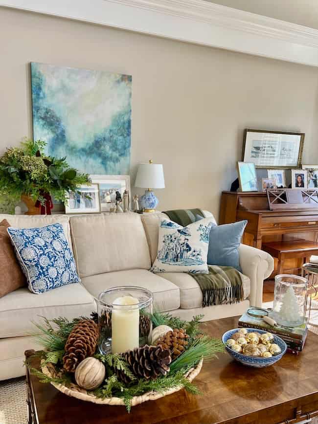A cozy living room with a beige sofa, blue and white pillows, and a green throw. A coffee table holds a candle centerpiece with pinecones and greenery. A piano and framed photos sit against the wall beneath abstract art.