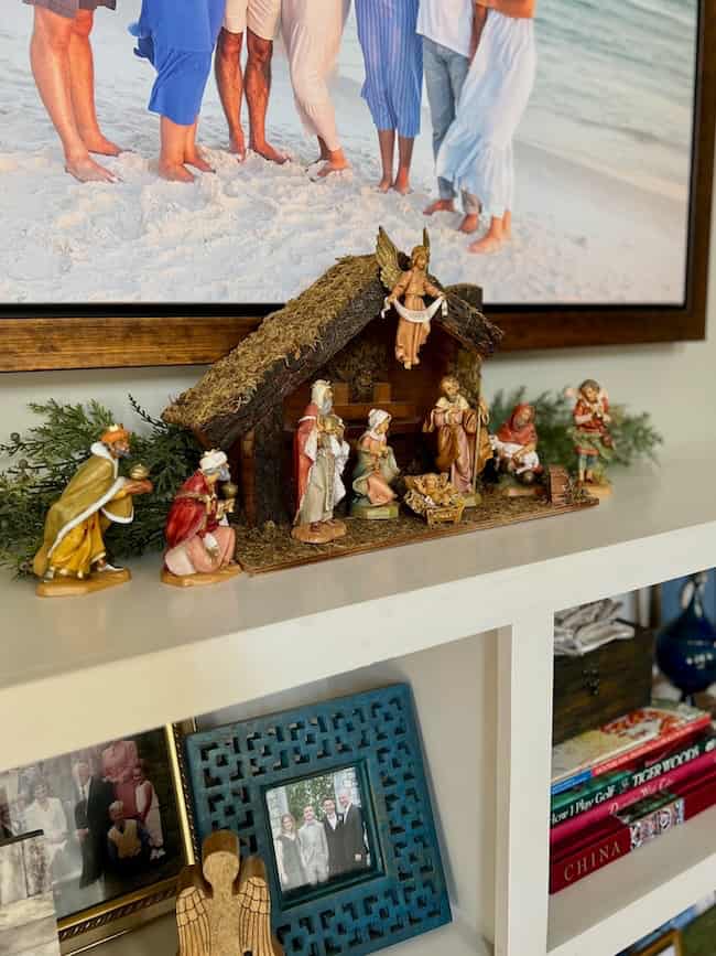 A nativity scene with figurines of Mary, Joseph, baby Jesus, the Wise Men, an angel, and animals is displayed on a white shelf beneath a framed beach family photo. Books and ornaments are visible on the shelves below.