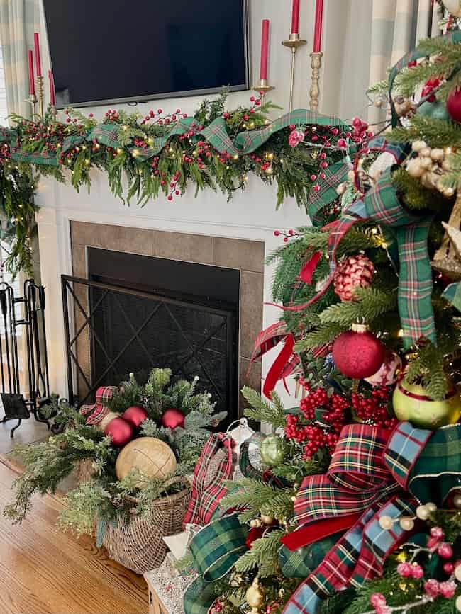 A festive living room with a decorated Christmas tree, plaid ribbon, red ornaments, greenery, and berries. The mantel above the fireplace is adorned with matching garland and red candles. A basket with ornaments sits by the hearth.