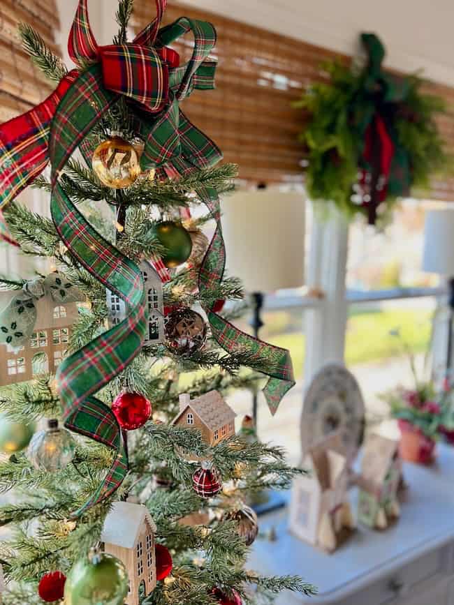 A small Christmas tree decorated with plaid ribbon, miniature houses, and ornaments sits by a window with bamboo blinds. Wrapped gifts and greenery with a bow are visible on a table and in the background.