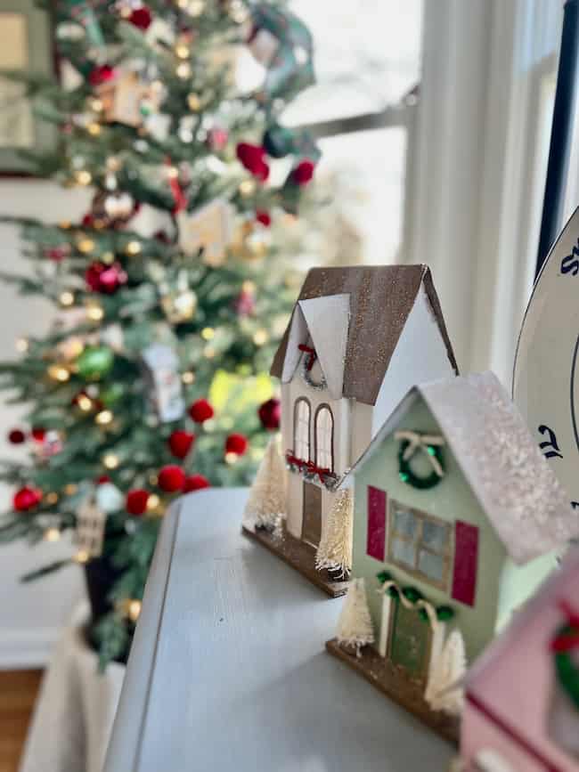 Two small decorative houses with holiday wreaths and snowy roofs sit on a table in the foreground. A Christmas tree with ornaments and lights is blurred in the background by a window.
