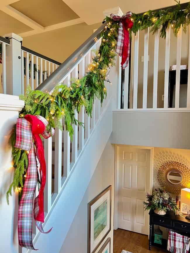 A staircase railing decorated with green garland, string lights, and large red plaid bows. The garland wraps around the banister in a bright, festive indoor setting with holiday decor visible below.